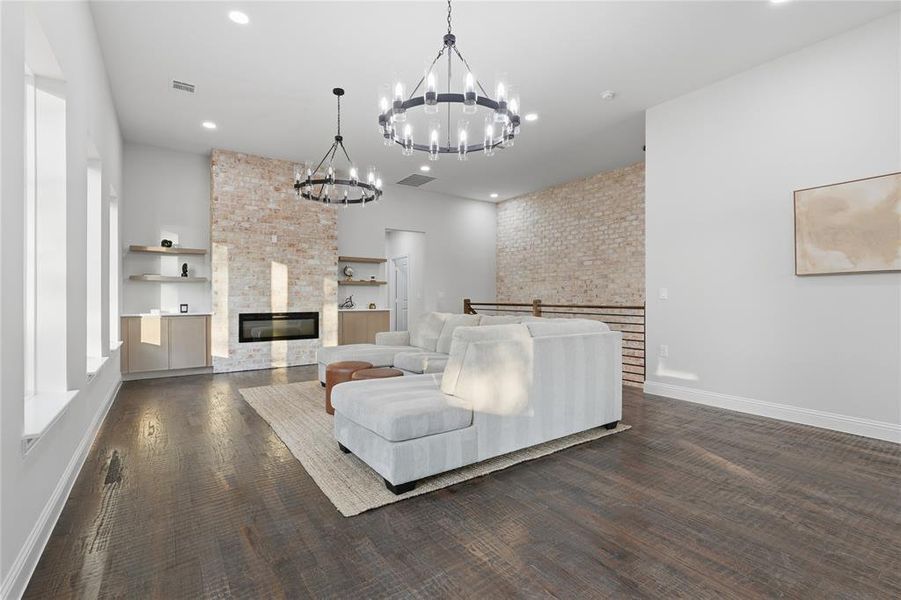 Spacious living area featuring a brick accent wall with a linear fireplace, custom built-in shelving, dark wood-finish flooring, and two contemporary chandeliers Spacious living area featuring a brick accent wall with a linear fireplace, custom built-in shelving, dark wood-finish flooring, and two contemporary chandeliers