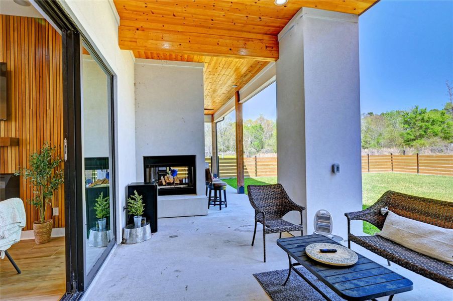 This photo showcases a modern outdoor patio with a cozy fireplace, covered by a wood-paneled ceiling. It features stylish seating and overlooks a spacious fenced backyard, perfect for relaxation and entertaining.