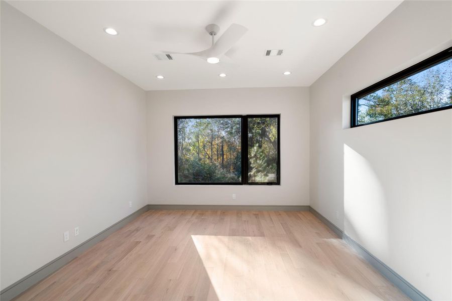 This room features sleek hardwood floors, large windows offering plenty of natural light and a view of nature, recessed lighting, and a modern ceiling fan, creating a bright and inviting space.