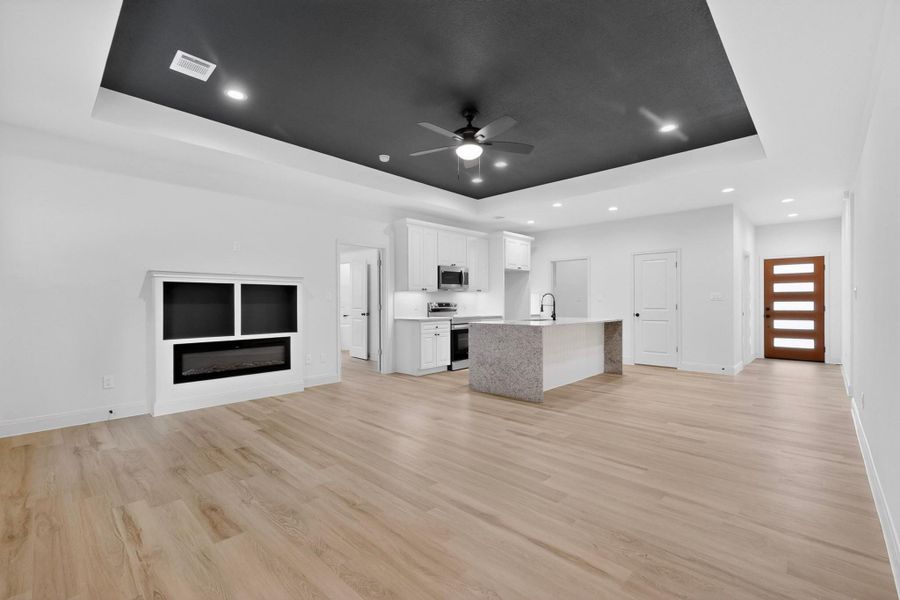 Open-concept living space featuring wood-finish flooring, a contemporary built-in fireplace, and a recessed ceiling with integrated lighting Open-concept living space featuring wood-finish flooring, a contemporary built-in fireplace, and a recessed ceiling with integrated lighting