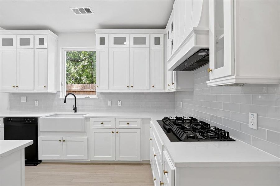 White shaker cabinetry with brass hardware, a white apron-front sink with matte black faucet, light grey subway tile backsplash, white countertops, and a built-in gas cooktop with matching range hood