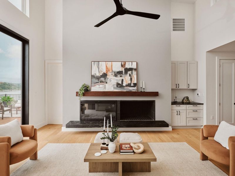 Spacious room featuring high ceilings, a grand fireplace with a dark stone hearth, a built-in wet bar with light cabinetry and dark countertops, wood-finish flooring, and expansive glass doors opening to an outdoor deck