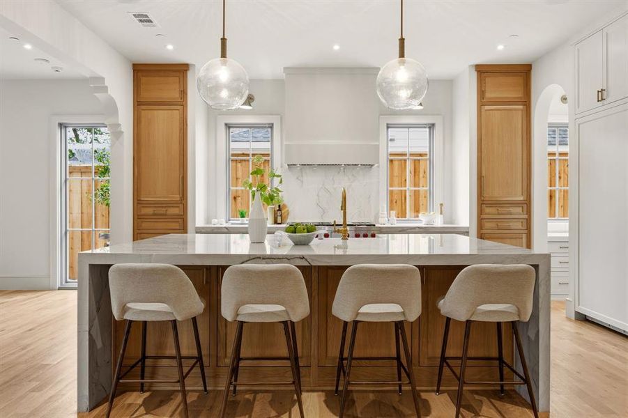Spacious kitchen featuring a large central island with seating, natural wood cabinetry, light-colored countertops, and hardwood flooring Spacious kitchen featuring a large central island with seating, natural wood cabinetry, light-colored countertops, and hardwood flooring