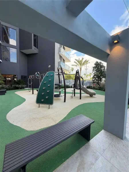 Outdoor recreation area featuring a multi-component play structure with a climbing wall, synthetic rubber surface, and a covered architectural pergola