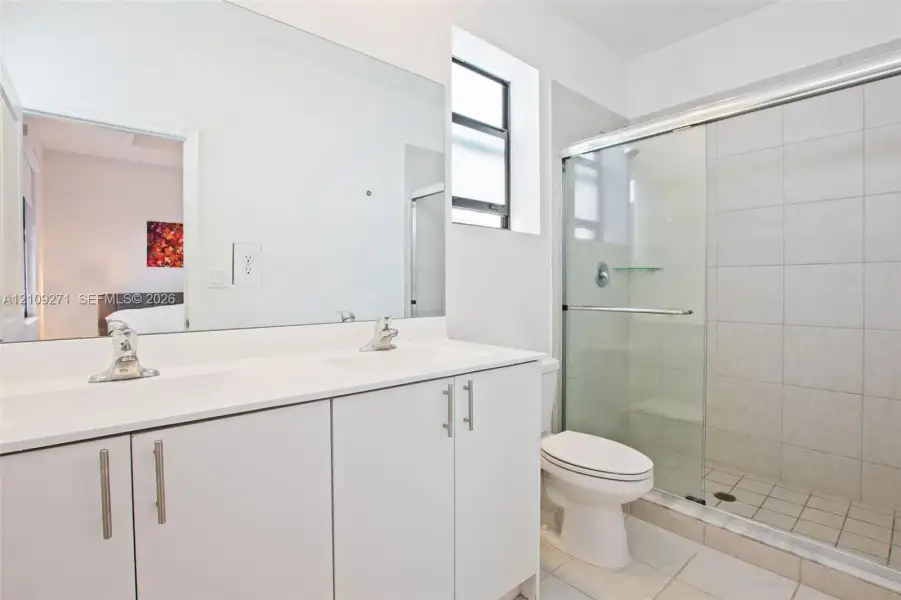 Dual-sink vanity with a solid-surface countertop, full-width mirror, glass-enclosed shower featuring tiled surround, and ceramic tile flooring