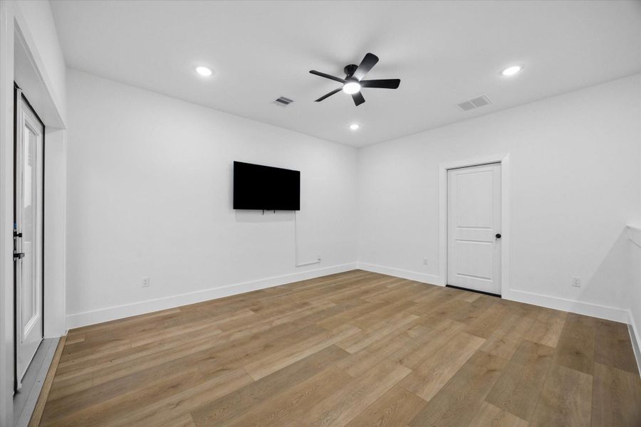 This room features sleek wooden flooring, a ceiling fan, recessed lighting, and a mounted TV. It offers a minimalist design with white walls and a door leading to another space, ideal for a modern living area or media room. This room features sleek wooden flooring, a ceiling fan, recessed lighting, and a mounted TV. It offers a minimalist design with white walls and a door leading to another space, ideal for a modern living area or media room.