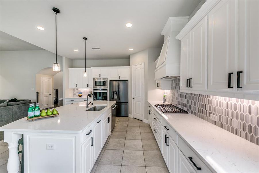 Kitchen featuring custom exhaust hood, backsplash, light tile floors, a kitchen island with sink, and appliances with stainless steel finishes