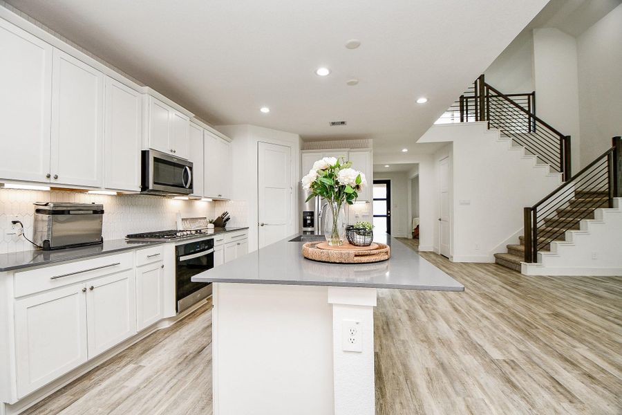 This photo features a modern kitchen with sleek white cabinetry, stainless steel appliances, and a large stylish island with a quartz countertop. The open layout extends to a staircase with modern horizontal iron railing, and the space is enhanced by bright lighting. This photo features a modern kitchen with sleek white cabinetry, stainless steel appliances, and a large stylish island with a quartz countertop. The open layout extends to a staircase with modern horizontal iron railing, and the space is enhanced by bright lighting.