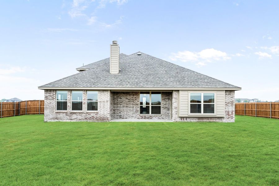 Rear view of single-story brick home with chimney, large windows, and fenced backyard with green lawn