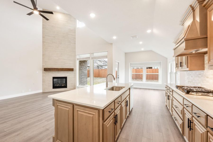Open kitchen with wood cabinets, center island with sink, and fireplace in adjoining living area with vaulted ceiling Open kitchen with wood cabinets, center island with sink, and fireplace in adjoining living area with vaulted ceiling