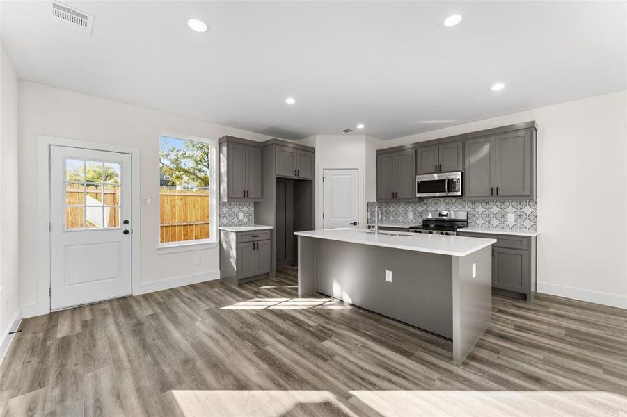 Kitchen with gray cabinets, stainless steel appliances, backsplash, a center island with sink, and light wood-type flooring Kitchen with gray cabinets, stainless steel appliances, backsplash, a center island with sink, and light wood-type flooring