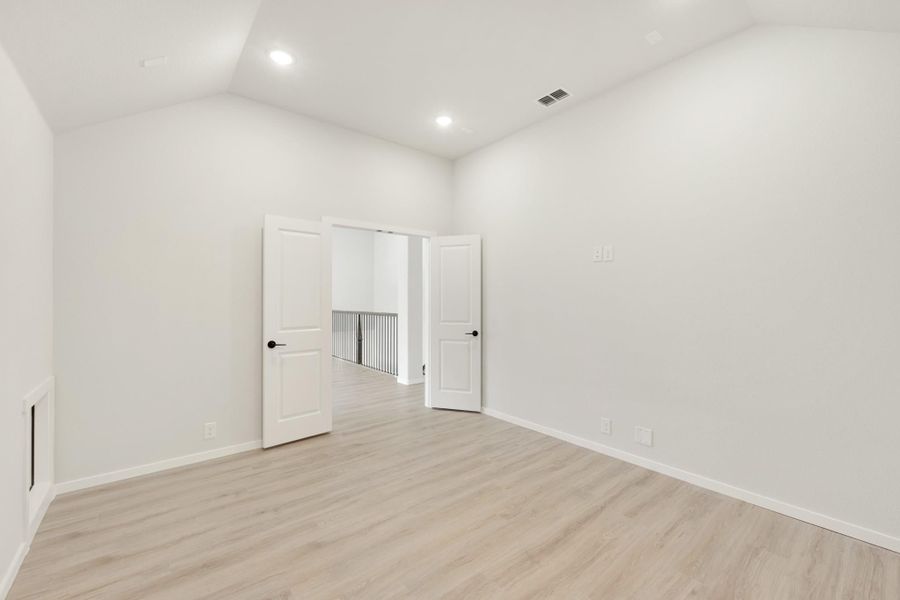 Empty bedroom with light wood floors, white walls, vaulted ceiling, and double doors opening to hallway.