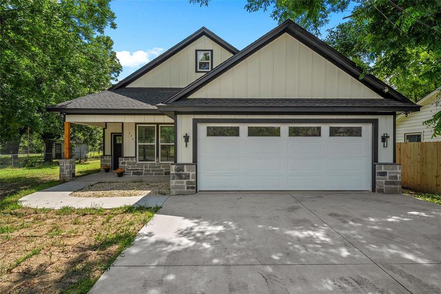 Contemporary exterior featuring light vertical siding, dark trim, a two-car garage with transom windows, stone veneer accents, and a covered front porch with a wood beam post Contemporary exterior featuring light vertical siding, dark trim, a two-car garage with transom windows, stone veneer accents, and a covered front porch with a wood beam post