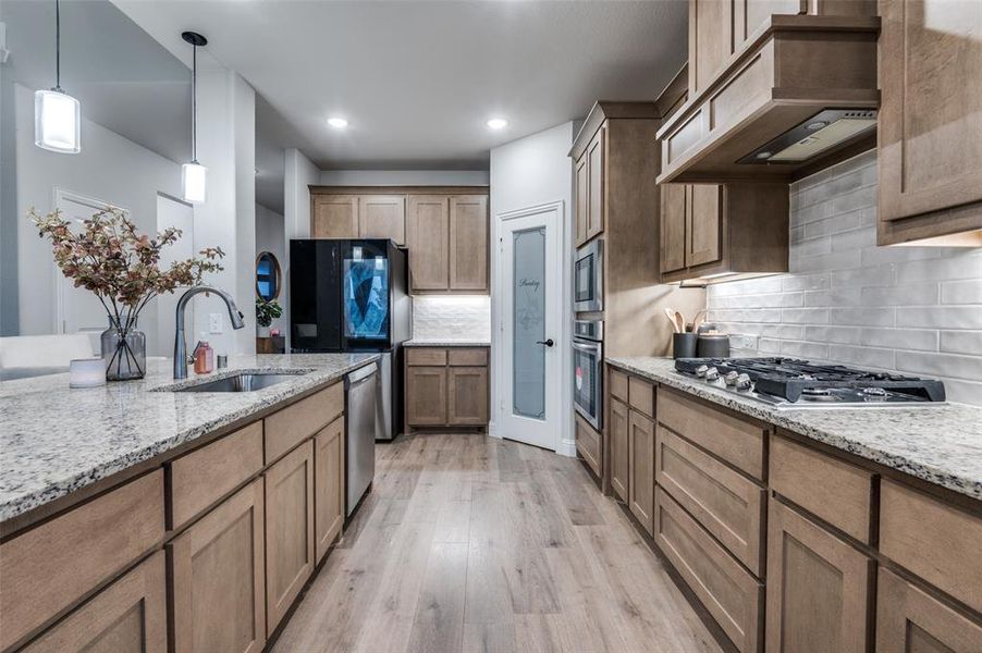 Kitchen featuring light wood-style flooring, light stone countertops, pendant lighting, wall chimney exhaust hood, and backsplash Kitchen featuring light wood-style flooring, light stone countertops, pendant lighting, wall chimney exhaust hood, and backsplash