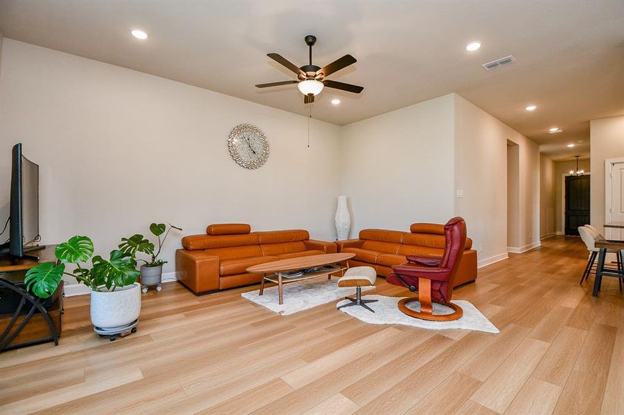 This spacious living room features modern, light wood flooring and neutral walls, providing a warm and inviting atmosphere. It includes stylish brown leather seating, a ceiling fan, recessed lighting, and a large TV, perfect for relaxation and entertainment.