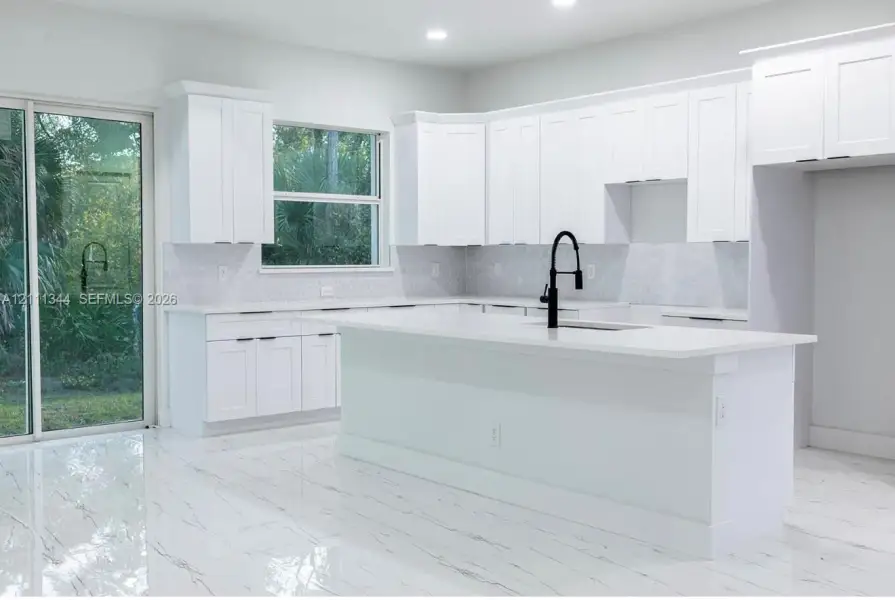 Kitchen featuring white Shaker-style cabinetry, a central island with a quartz countertop, geometric pattern backsplash, polished stone-finish flooring, and a matte black gooseneck faucet Kitchen featuring white Shaker-style cabinetry, a central island with a quartz countertop, geometric pattern backsplash, polished stone-finish flooring, and a matte black gooseneck faucet