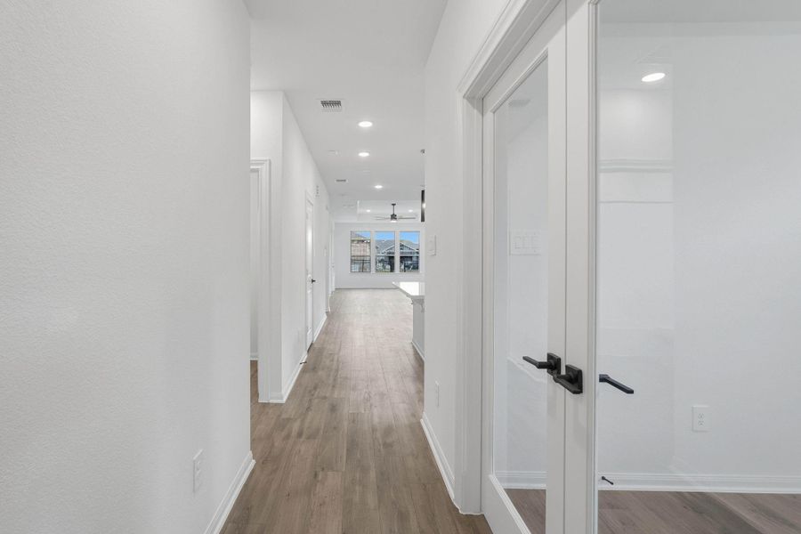 The hallway draws you in with warm floors and soft lighting, opening toward the main living area while glass doors hint at flexible spaces along the way.