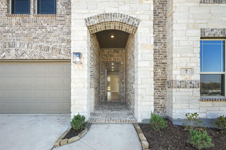 Home entrance with arched brick doorway, stone exterior, wall sconce light, and two-car garage door nearby. Home entrance with arched brick doorway, stone exterior, wall sconce light, and two-car garage door nearby.