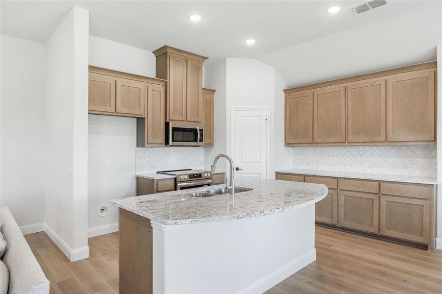 Kitchen with light wood cabinets, granite island with sink, stainless appliances, and herringbone tile backsplash