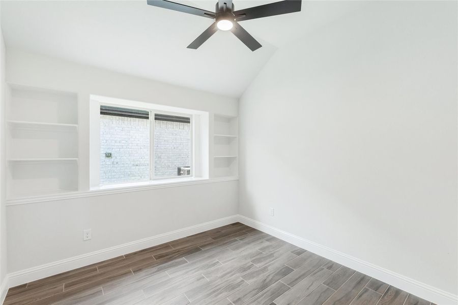 Empty room with vaulted ceiling, ceiling fan, built-in shelves, and wood-look tile floors Empty room with vaulted ceiling, ceiling fan, built-in shelves, and wood-look tile floors