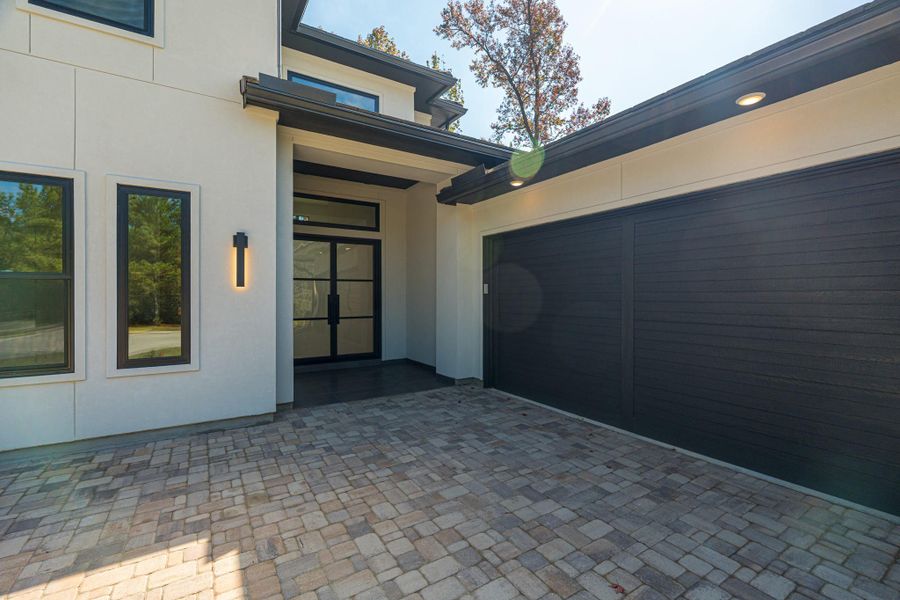 This modern home features a sleek exterior with a combination of large windows and a dark garage door. The entrance is inviting, with a covered entryway and stylish lighting. The paved driveway adds a sophisticated touch to the overall design.