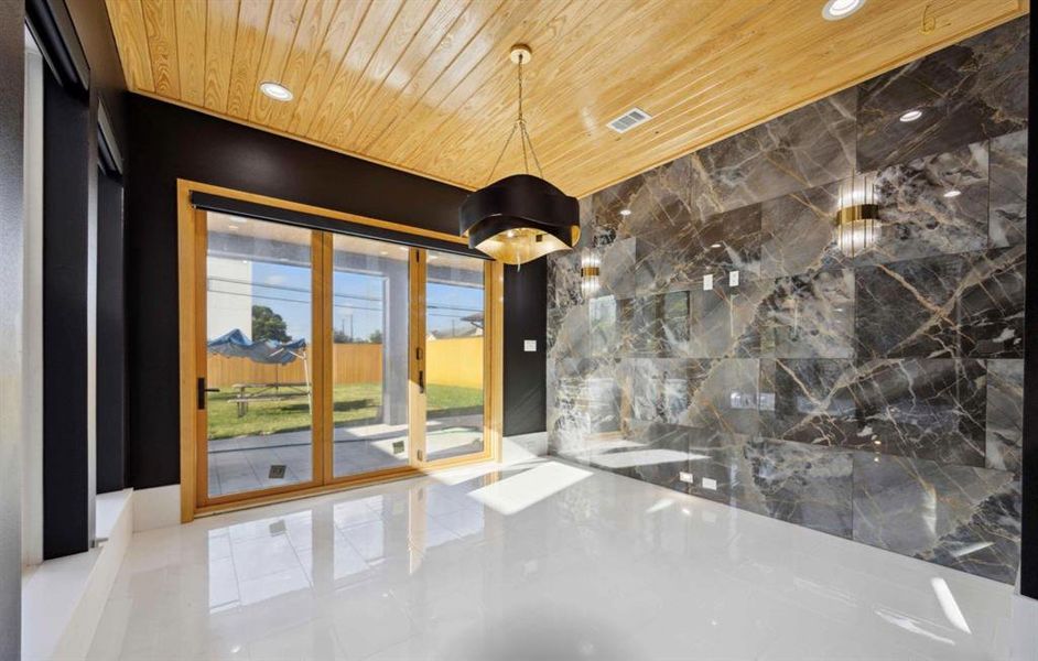 This contemporary dining room space features polished white floors, a striking marble wall, and a wood-paneled ceiling, complemented by sleek black trim and modern lighting. Large glass doors offer abundant natural light and a view of the yard.