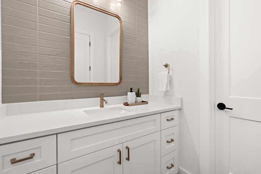 This bathroom features a modern design with a sleek white vanity, quartz countertop, and elegant brass fixtures. The taupe tiled backsplash adds a touch of sophistication, complemented by a large framed mirror.