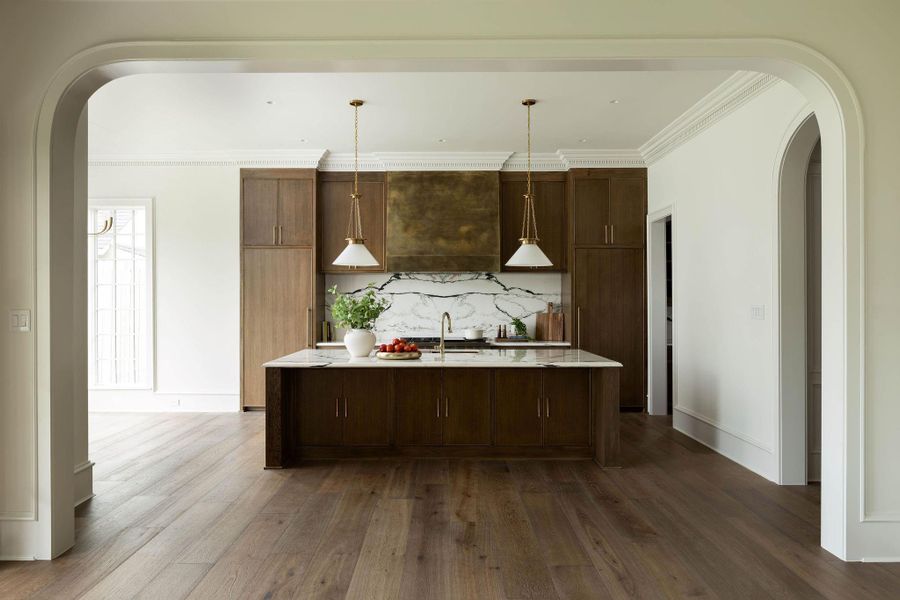 Chef's kitchen featuring custom solid wood cabinetry with brass hardware, a large center island with marble countertop and undermount brass faucet, full-height dramatic marble slab backsplash, and an oversized antiqued brass range hood. Detailed dentil crown molding and wide-plank hardwood floors throughout. Chef's kitchen featuring custom solid wood cabinetry with brass hardware, a large center island with marble countertop and undermount brass faucet, full-height dramatic marble slab backsplash, and an oversized antiqued brass range hood. Detailed dentil crown molding and wide-plank hardwood floors throughout.