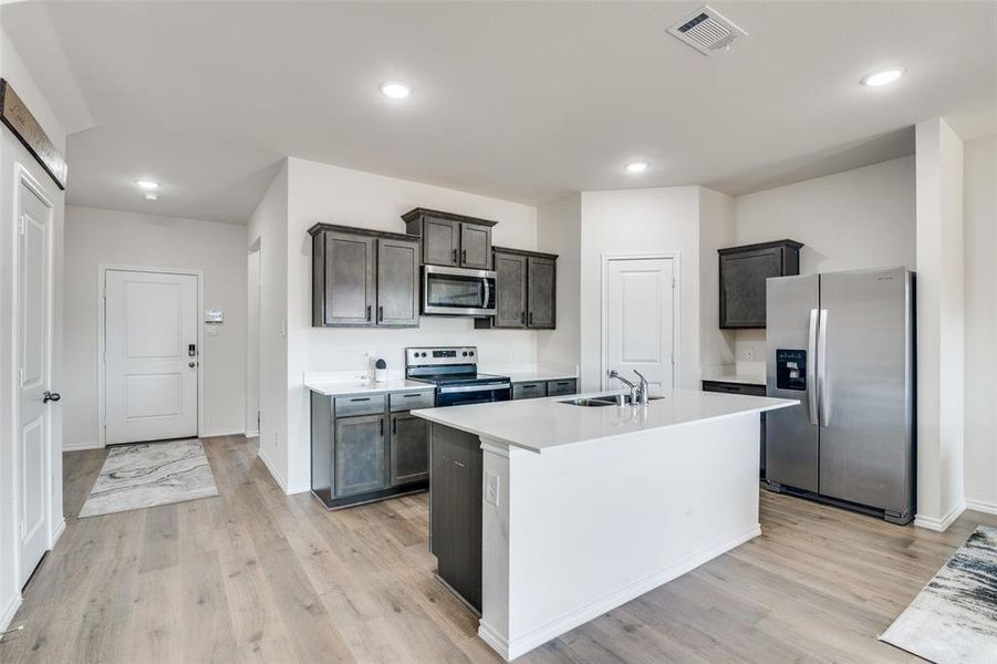 Kitchen featuring stainless steel appliances, light wood-style flooring, an island with sink, recessed lighting, and dark wood finish cabinetry Kitchen featuring stainless steel appliances, light wood-style flooring, an island with sink, recessed lighting, and dark wood finish cabinetry