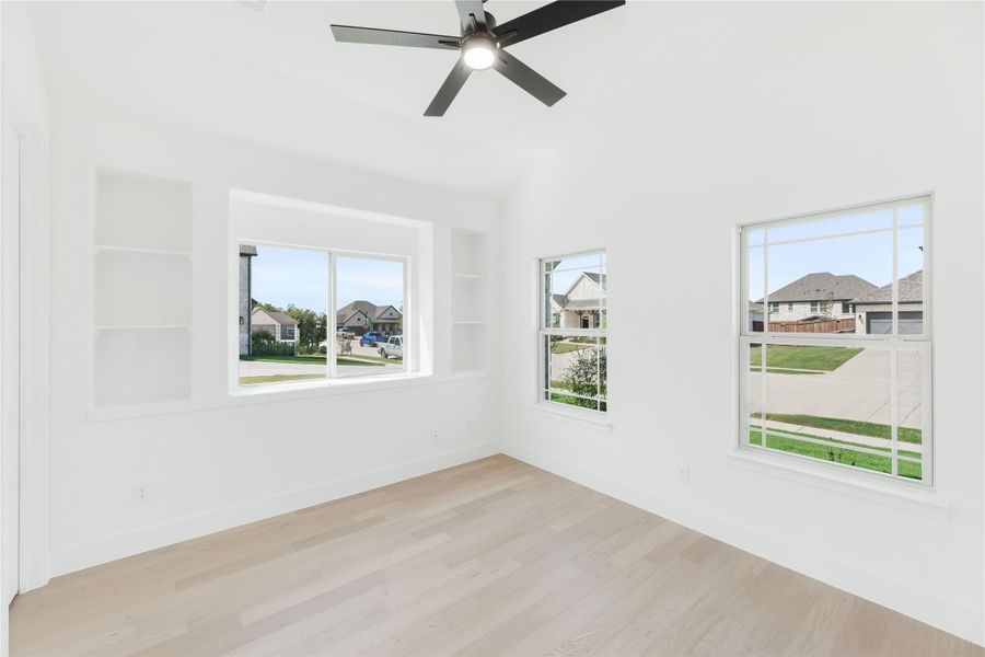 Bright empty bedroom with hardwood floors, ceiling fan, built-in shelves, and three windows overlooking a neighborhood