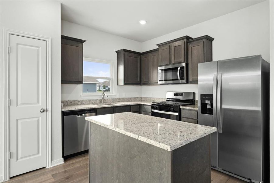 Kitchen featuring stainless steel appliances, dark brown cabinets, light stone counters, dark wood finished floors, and a center island