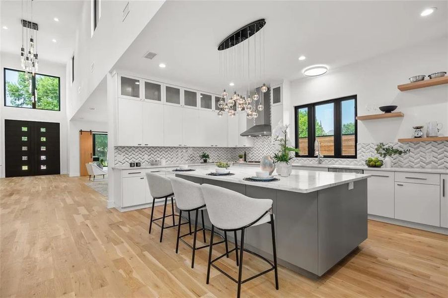 Open-concept kitchen with a large central island, light wood-finish flooring, white cabinetry with glass upper doors, a chevron pattern tile backsplash, and contemporary pendant lighting Open-concept kitchen with a large central island, light wood-finish flooring, white cabinetry with glass upper doors, a chevron pattern tile backsplash, and contemporary pendant lighting
