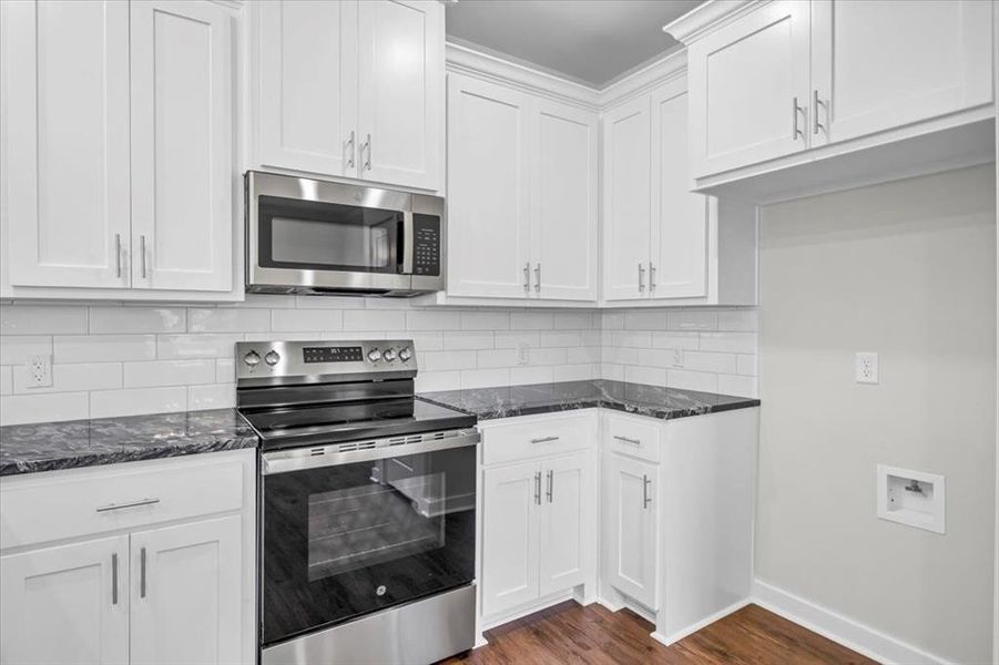 Kitchen with stainless steel appliances, dark stone countertops, dark wood-type flooring, backsplash, and white cabinetry