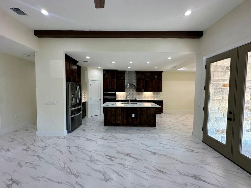 Kitchen with a center island with sink, decorative backsplash, dark brown cabinets, stainless steel fridge, and light marble finish flooring