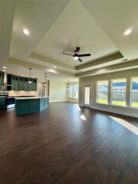 This open-concept living space features a modern kitchen with green cabinetry, a large island, and stylish pendant lighting. The room is accentuated by a tray ceiling with recessed lights and a ceiling fan. Expansive windows provide ample natural light, and the dark wood flooring adds a touch of elegance. This open-concept living space features a modern kitchen with green cabinetry, a large island, and stylish pendant lighting. The room is accentuated by a tray ceiling with recessed lights and a ceiling fan. Expansive windows provide ample natural light, and the dark wood flooring adds a touch of elegance.