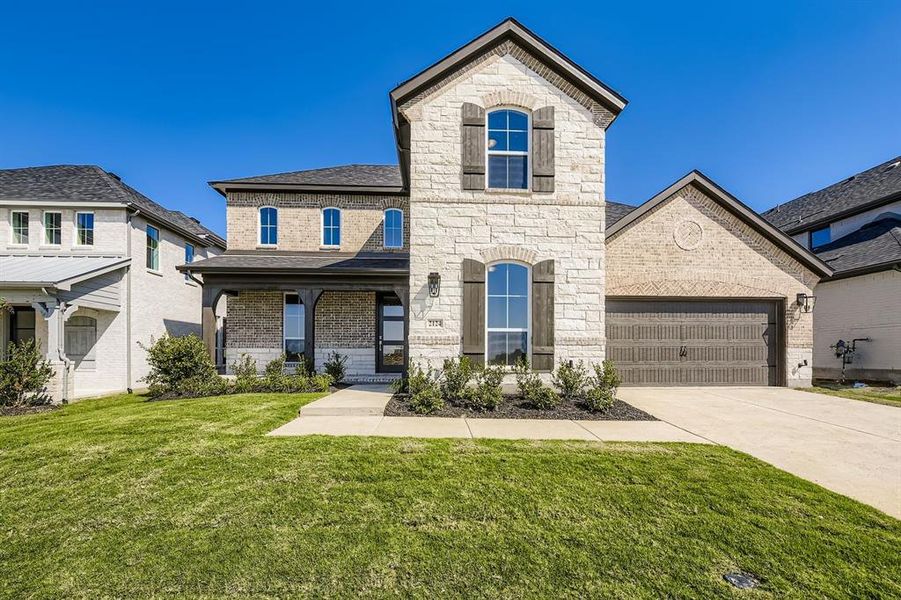 French country inspired facade with brick siding, stone siding, a front yard, covered porch, and driveway