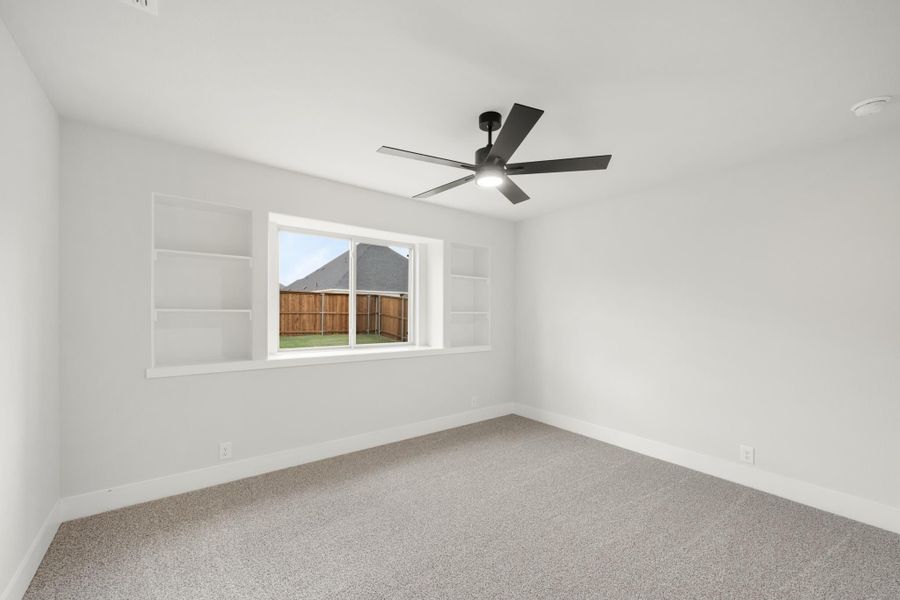 Empty bedroom with built-in shelves, ceiling fan, carpet flooring, and a window overlooking a fenced backyard