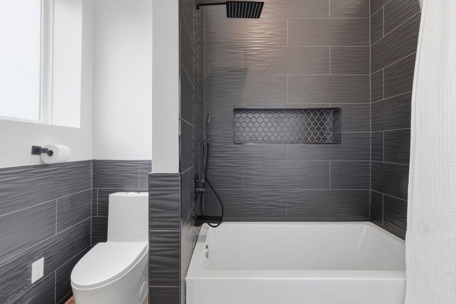 This bathroom features modern dark gray tile, a sleek white bathtub with a rainfall showerhead, and a compact toilet. A built-in shower niche offers convenient storage, and a window provides natural light.