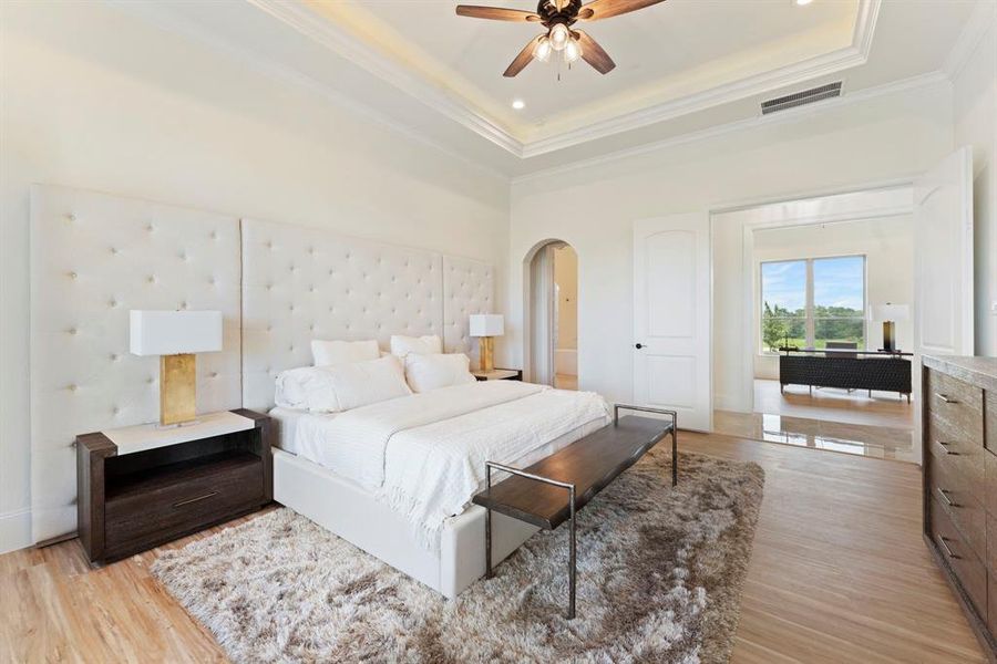 This is a spacious and elegantly appointed bedroom featuring a large tufted headboard, hardwood floors, a plush area rug, and high ceilings with a ceiling fan. Natural light streams in through a large window, offering a view of the outdoors. The room includes modern furnishings and a serene color palette, creating a luxurious and restful environment.