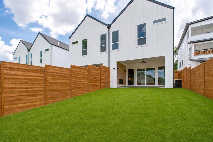 Expansive backyard featuring a manicured artificial turf lawn, a covered patio with a ceiling fan, and sliding glass doors