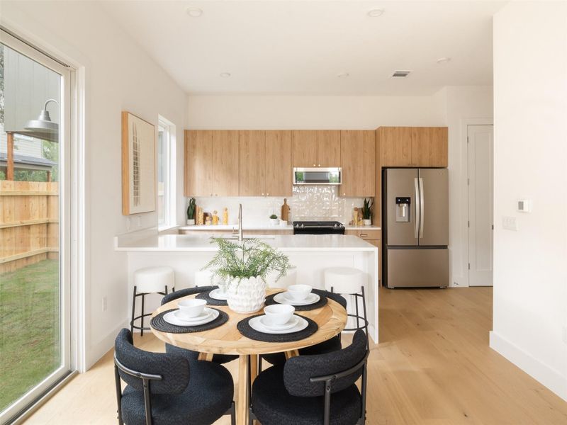 Open-concept kitchen and dining area featuring light wood-finish flooring, a white kitchen island with counter seating, light wood cabinetry, stainless steel appliances, and a tiled backsplash