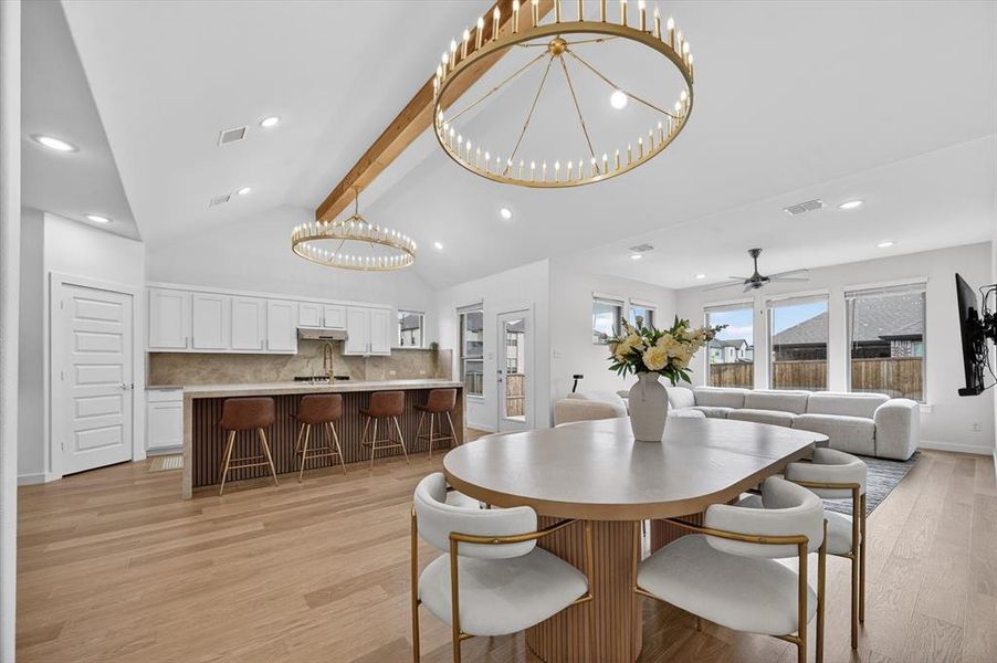 Open-concept living area featuring a vaulted ceiling with exposed wood beam, light wood-finish flooring, two large circular chandeliers, and a kitchen island with seating