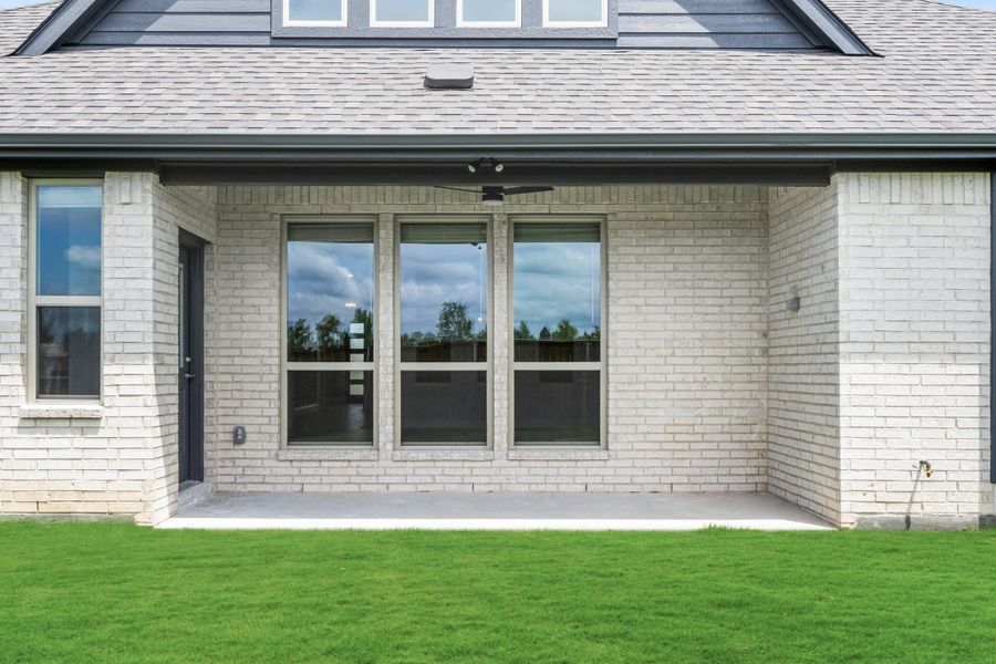 Rear exterior of brick home with covered patio, ceiling fan, large windows, and green lawn