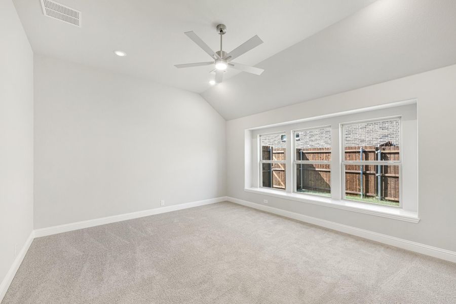 Empty bedroom with vaulted ceiling, ceiling fan, carpet flooring, and triple window overlooking backyard fence