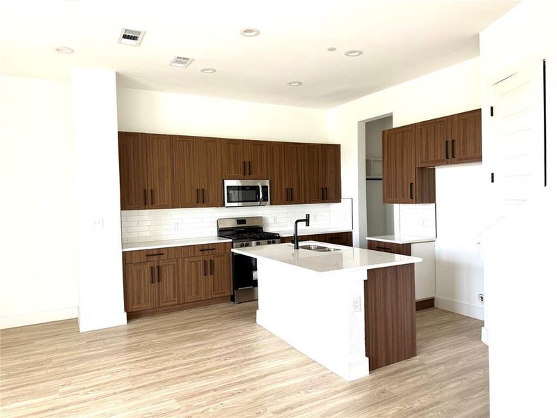 Kitchen featuring appliances with stainless steel finishes, light countertops, light wood-style floors, decorative backsplash, and a center island with sink