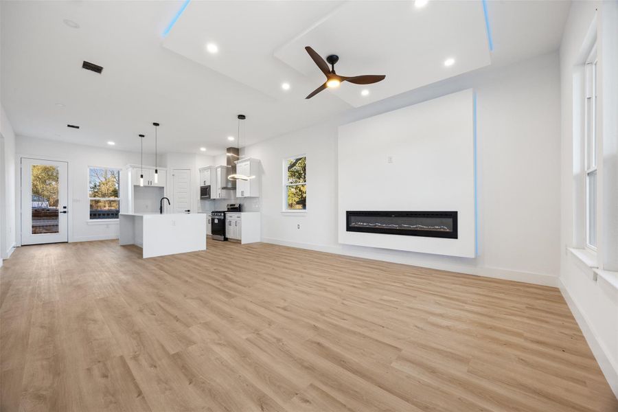 This modern open-concept living space features light wood flooring, a sleek electric fireplace, and a ceiling fan. The adjacent kitchen includes a large island, pendant lighting, and stainless steel appliances. Large windows and glass doors provide ample natural light.