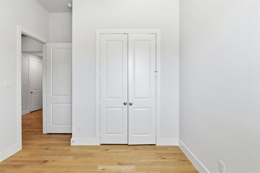 Interior room featuring light wood-finish flooring, white walls, white baseboards, and a white bi-fold closet door with brushed nickel hardware Interior room featuring light wood-finish flooring, white walls, white baseboards, and a white bi-fold closet door with brushed nickel hardware