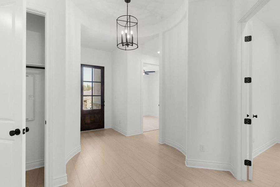 A graceful foyer with light wood flooring leads to a robust wooden entry door, bathed in the glow of a modern chandelier.