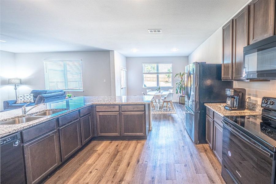 Kitchen with dark brown cabinets, black appliances, light wood-style floors, open floor plan, and light stone counters