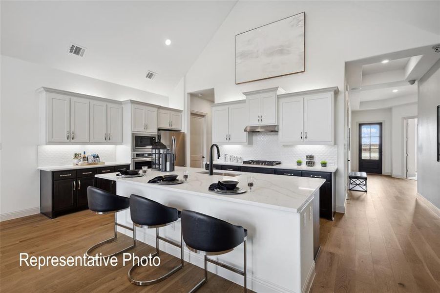 Open-concept kitchen featuring a large central island with seating, a high vaulted ceiling, two-tone cabinetry, wood-finish flooring, and a stainless steel appliance suite